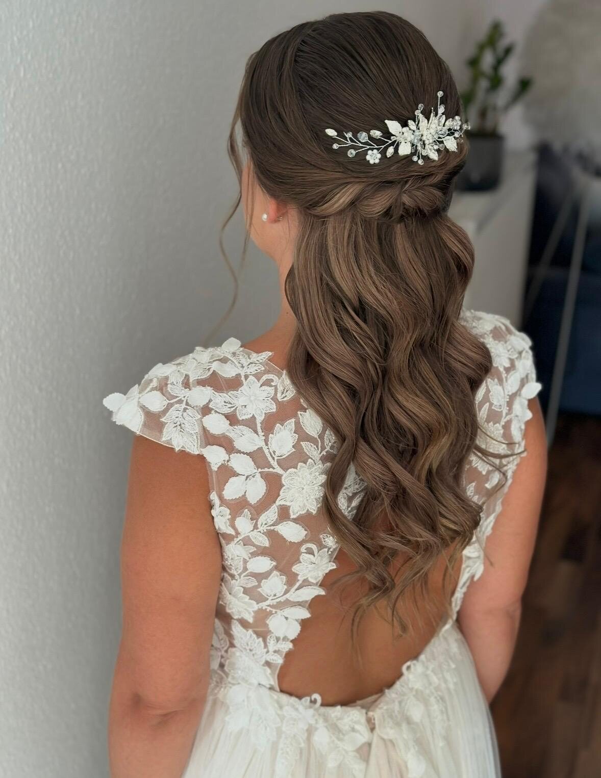 Braut von hinten mit langen, offenen Wellen und feinem floralem Haarschmuck in einem Spitzenkleid mit Flügelärmeln