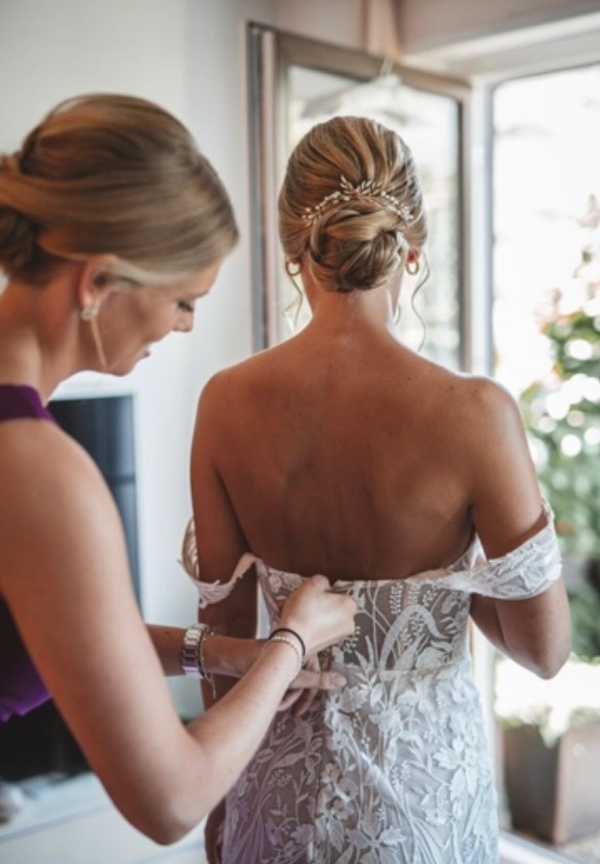 Braut wird beim Getting Ready in ihr Spitzenkleid geknöpft, geflochtene Hochsteckfrisur mit Haarschmuck
