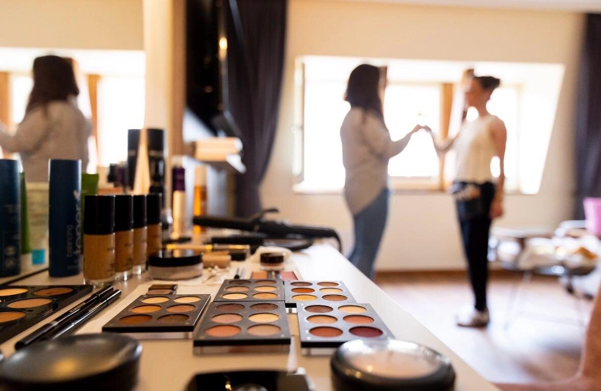 Make-up-Paletten und Pinsel im Vordergrund, Braut und Fotografin im Hintergrund am Fenster