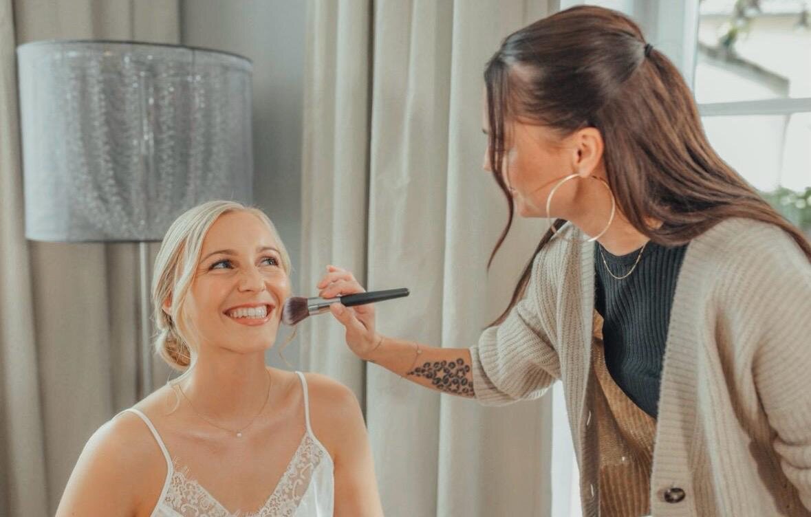 Stylistin trägt mit einem Pinsel Make-up bei einer lachenden Braut auf, helles Zimmer